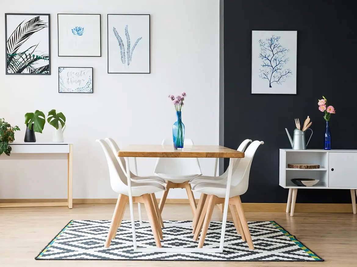 A modern dining area in Ulcinj, Montenegro real estate properties, featuring a wooden table, four white chairs, and a blue vase with flowers. A black-and-white rug covers the floor beneath framed botanical prints and minimal decorations by ekosphere.