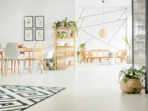 Bright, modern living and dining area in Ulcinj, Montenegro real estate properties, featuring light wood furniture, green plants, geometric wall art, a black and white patterned rug, and wicker chairs&mdash;all set against white walls and flooring.