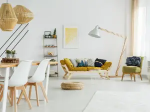 A modern living and dining area in Ulcinj, Montenegro, features white walls, wooden furniture, a yellow sofa with cushions, a green armchair, a floor lamp, decorative shelving, pendant lights, and a round woven rug&mdash;ideal for real estate properties seekers.