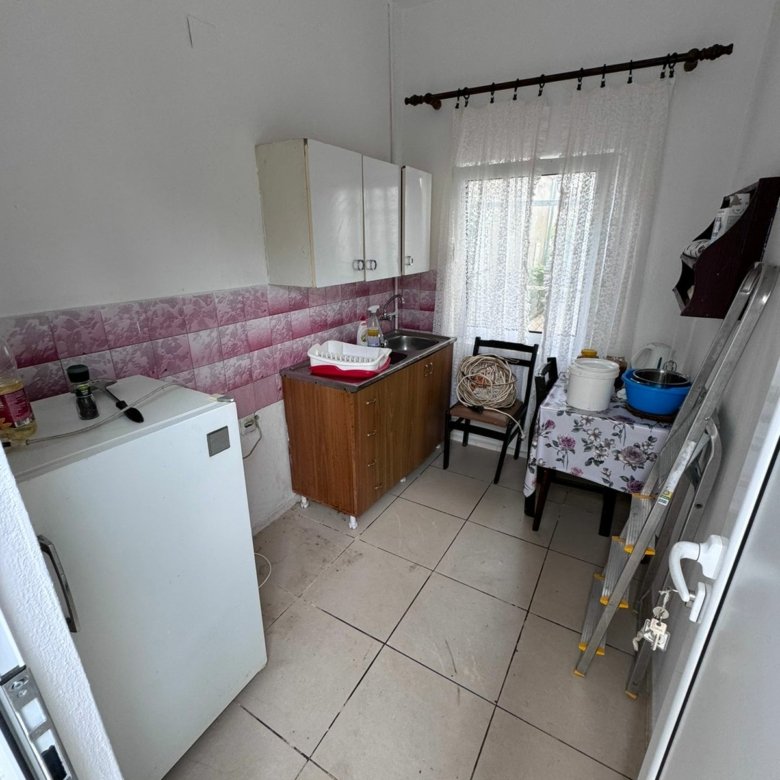 Cozy Ulcinj kitchen for sale: white tile floor, pink/white backsplash, wood cabinets, lace curtains. Exclusive at immorenta.me by ekosphere doo.