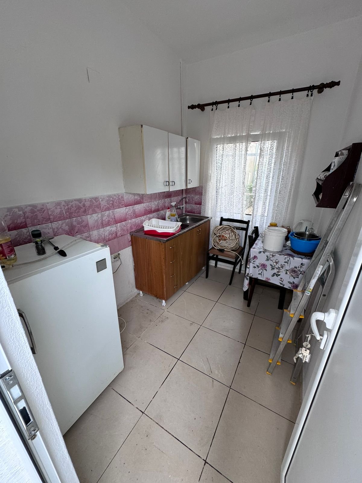 Cozy Ulcinj kitchen for sale: white tile floor, pink/white backsplash, wood cabinets, lace curtains. Exclusive at immorenta.me by ekosphere doo.
