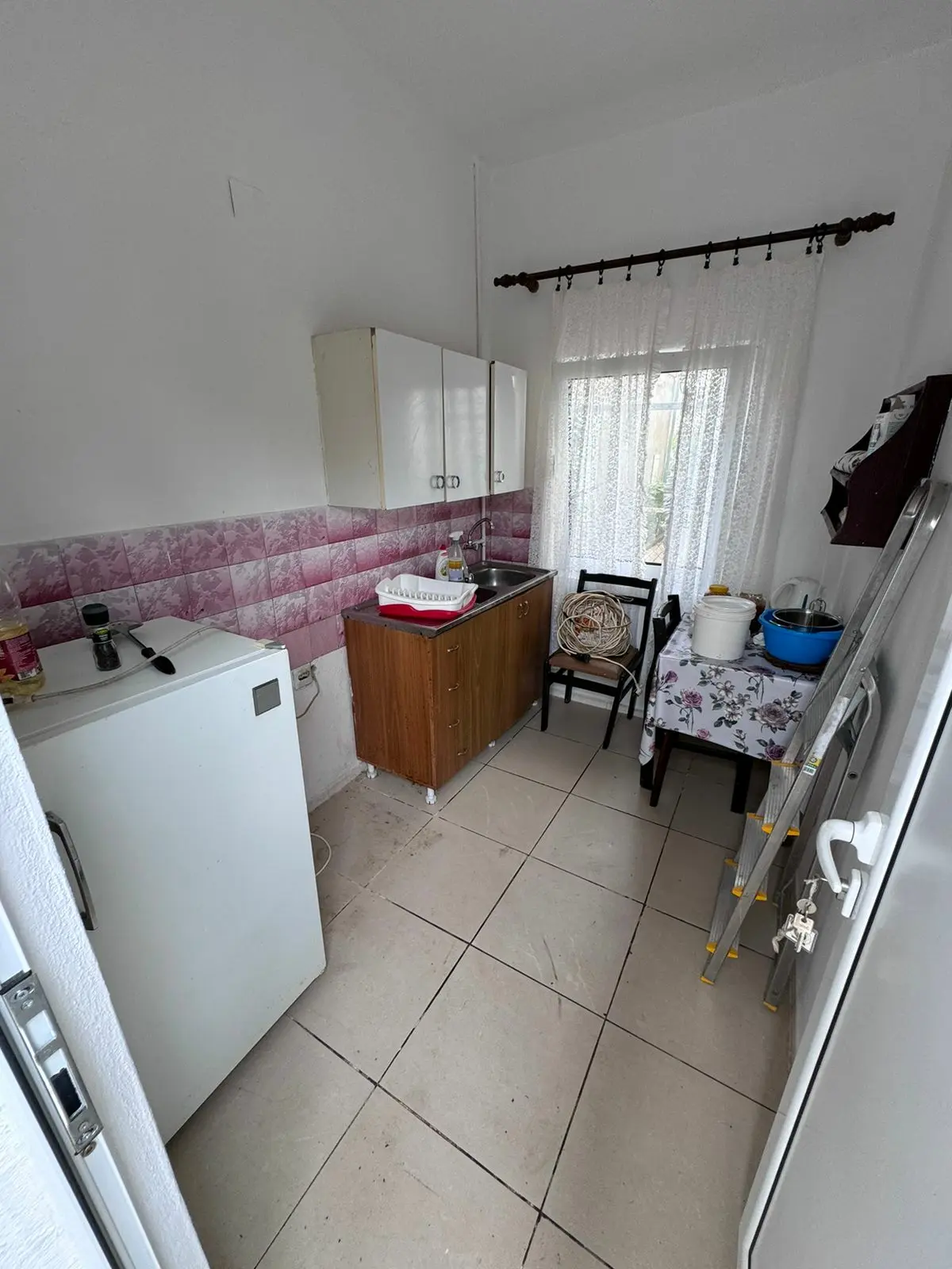 Cozy Ulcinj kitchen for sale: white tile floor, pink/white backsplash, wood cabinets, lace curtains. Exclusive at immorenta.me by ekosphere doo.