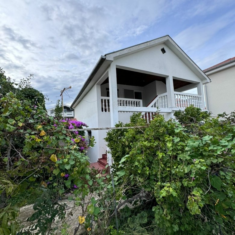 Elegant two-story white house with balcony, lush greenery, and purple flowers&mdash;prime Ulcinj real estate by immorenta.me.