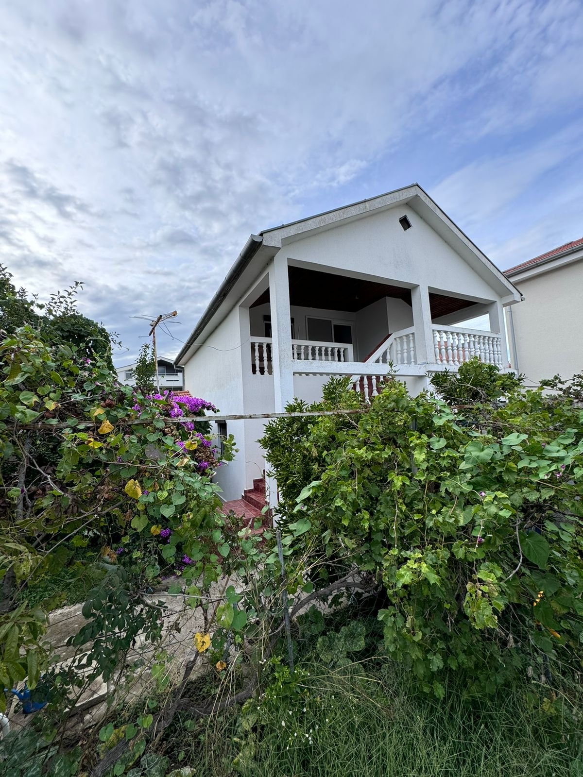 Elegant two-story white house with balcony, lush greenery, and purple flowers&mdash;prime Ulcinj real estate by immorenta.me.