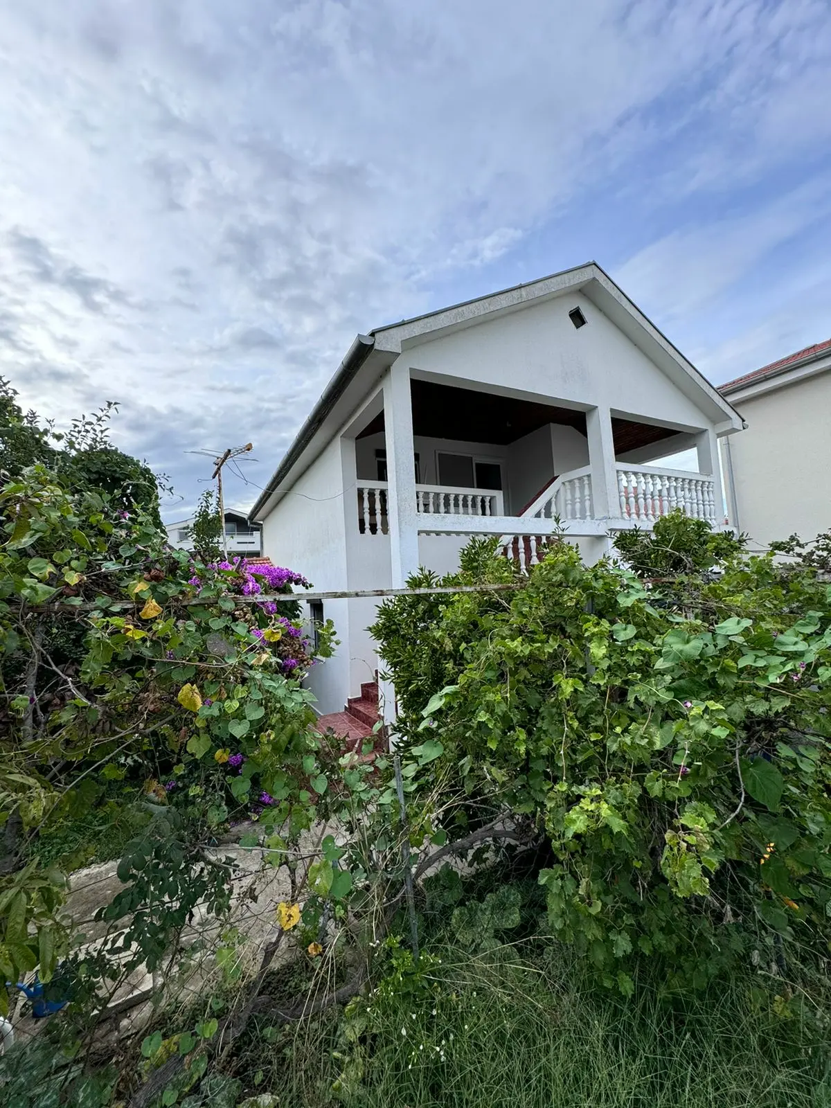 Elegant two-story white house with balcony, lush greenery, and purple flowers&mdash;prime Ulcinj real estate by immorenta.me.