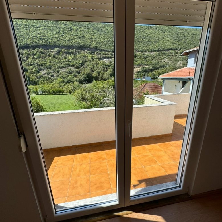Modern Ulcinj property: glass doors open to tiled balcony, scenic green hills, red-roof views; ideal real estate at immorenta.me.