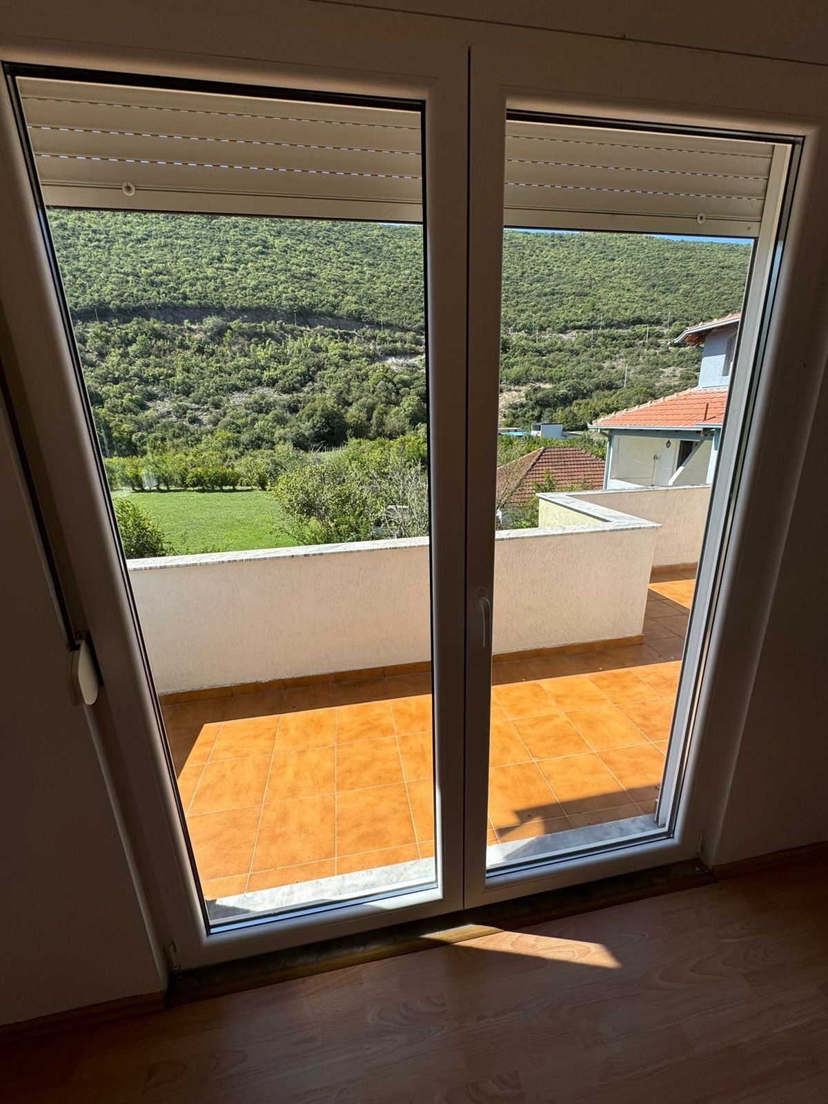 Modern Ulcinj property: glass doors open to tiled balcony, scenic green hills, red-roof views; ideal real estate at immorenta.me.