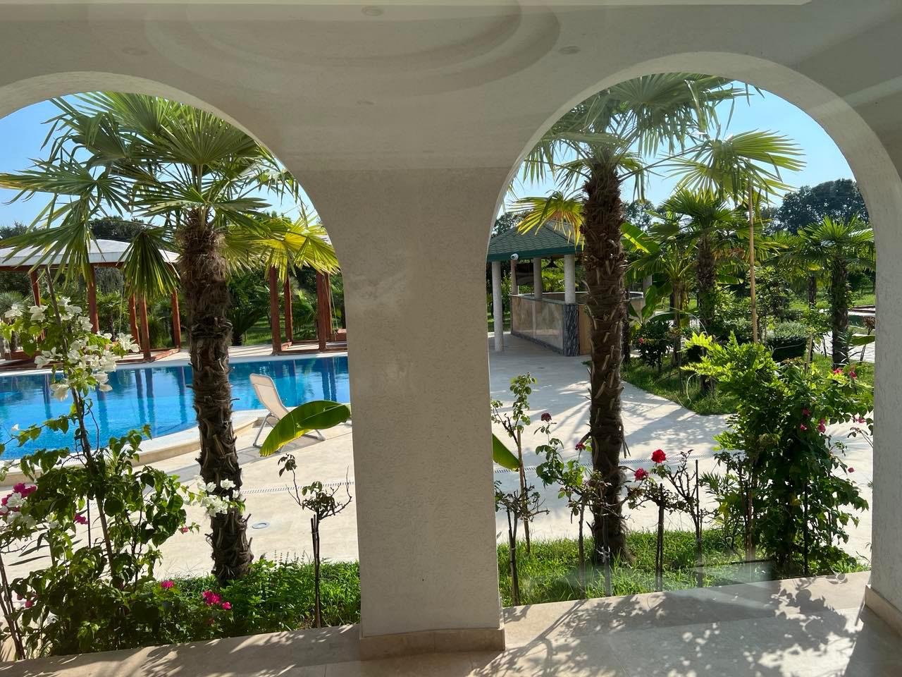 Stunning Ulcinj villa for sale: tropical garden, palm trees, pool view through white arches. Invest via immorenta.me by Ekosphere Doo.