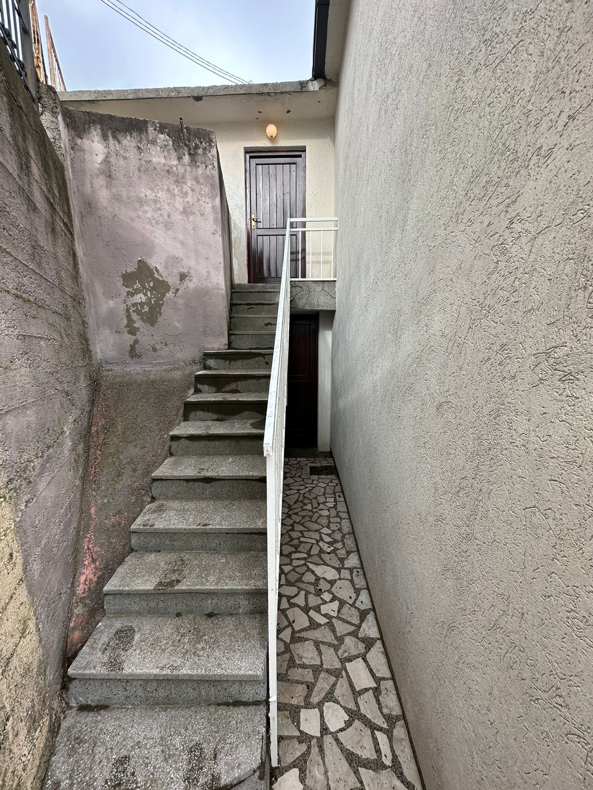 Prime Ulcinj property: concrete stairs, white railing, dual dark brown doors, stone path, and weathered fa&ccedil;ade. Invest via immorenta.me by ekosphere doo.