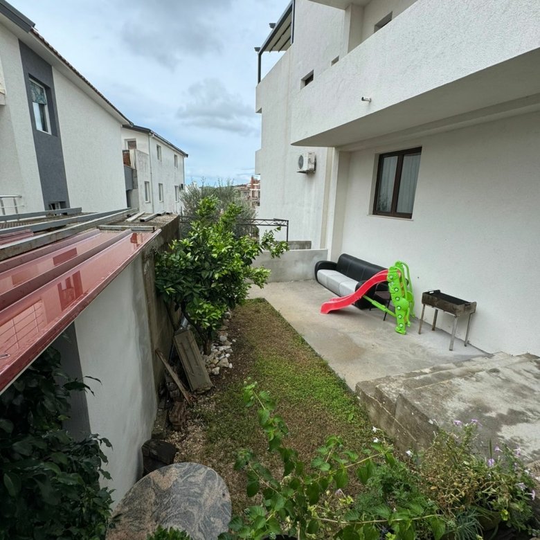 Charming Ulcinj home for sale: grassy backyard, potted plants, patio with sofa, grill, kids&rsquo; slide. Montenegro-listings.me by Ekosphere.