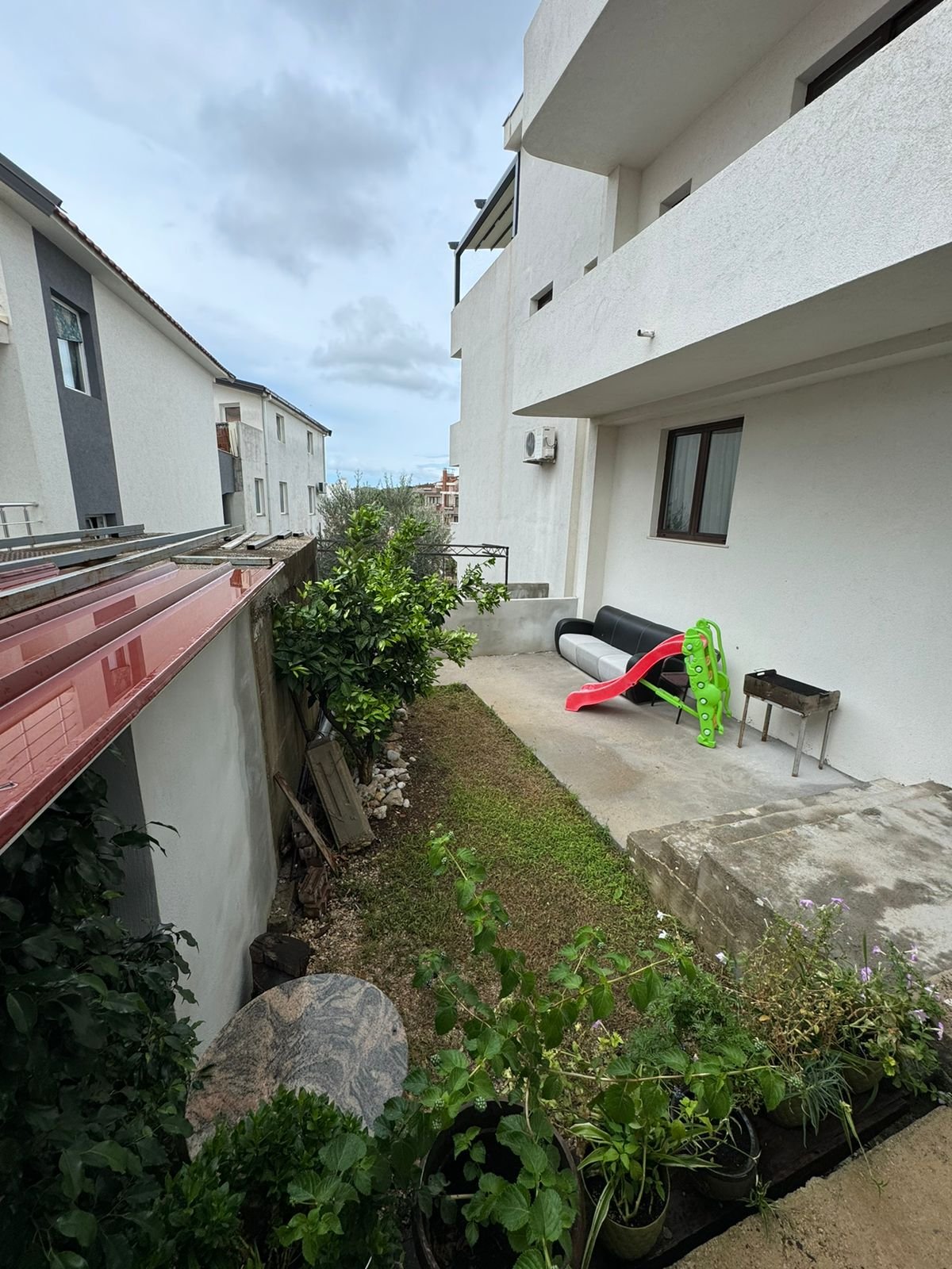 Charming Ulcinj home for sale: grassy backyard, potted plants, patio with sofa, grill, kids&rsquo; slide. Montenegro-listings.me by Ekosphere.