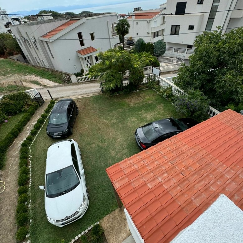 Aerial view of Ulcinj real estate: red-roofed home, grassy lot with two cars, white houses nearby&mdash;your investment at immorenta.me by ekosphere doo.
