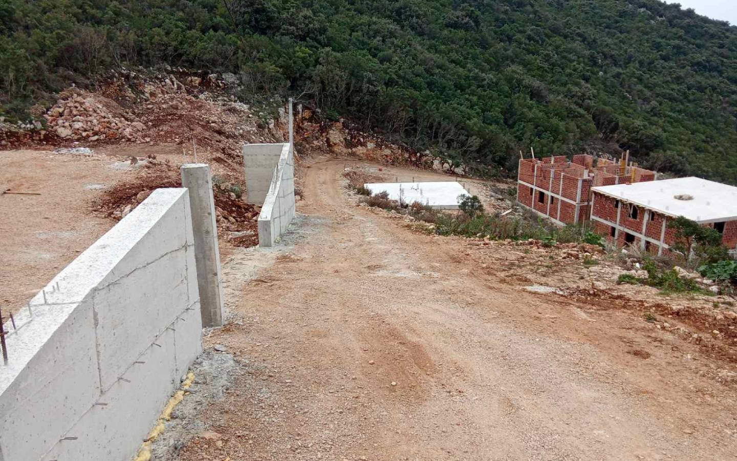 Prime investment opportunity in Ulcinj: sloping access, concrete wall, and unfinished brick homes with lush hillside views. Montenegro-listings.me by ekosphere doo.