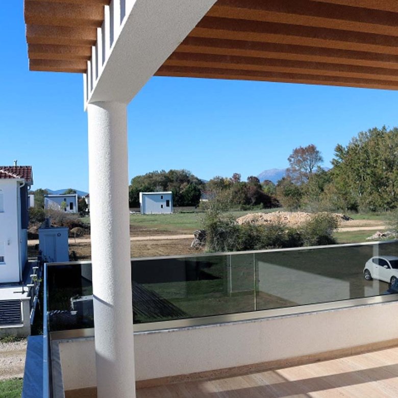 Discover tranquil Ulcinj living from Adana Villa&rsquo;s covered balcony&mdash;glass railings reveal lush land, serene homes, and blue skies. Invest with immorenta.me by ekosphere doo.
