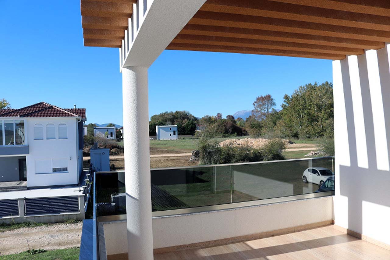 Discover tranquil Ulcinj living from Adana Villa&rsquo;s covered balcony&mdash;glass railings reveal lush land, serene homes, and blue skies. Invest with immorenta.me by ekosphere doo.