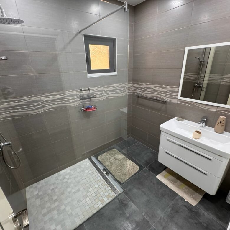 Stylish Ulcinj bathroom: gray tiles, walk-in shower, rainfall heads, vanity, mirror&mdash;premium real estate by immorenta.me.
