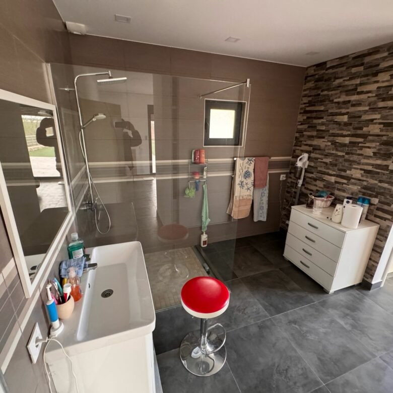 Modern Ulcinj bathroom with walk-in shower, stylish sink, red stool, and stone-tile walls. Exclusive on immorenta.me by ekosphere doo.