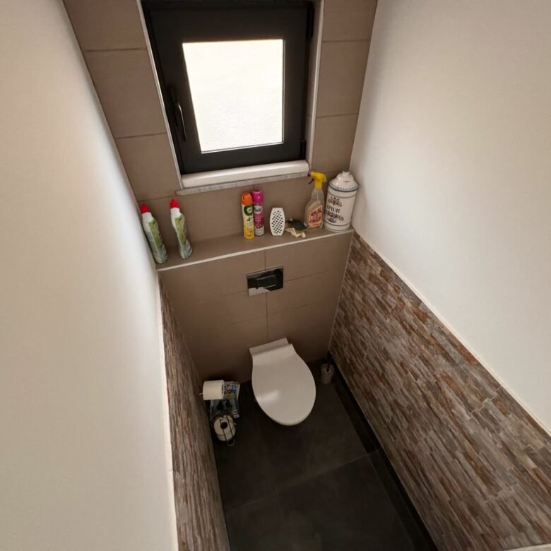Modern small bathroom in Ulcinj with wall-mounted toilet, eco-friendly brown tiles, and window. Featured on immorenta.me by ekosphere doo.