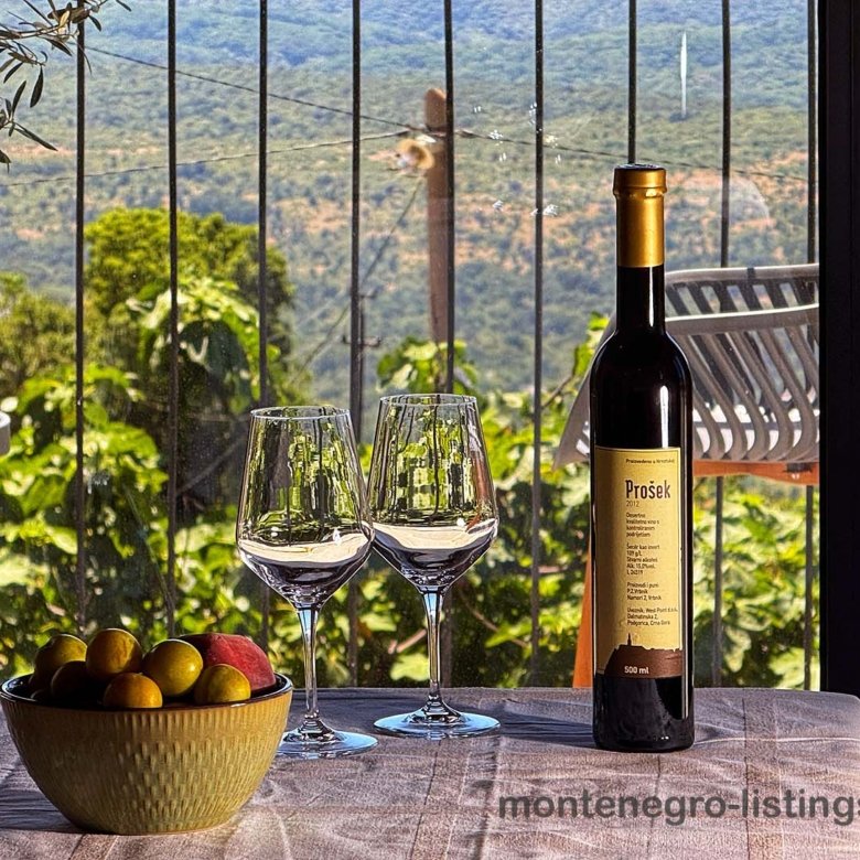 Pro&scaron;ek wine, glasses, and fresh fruit set by a window with green hill views&mdash;experience Ulcinj real estate luxury with immorenta.me.