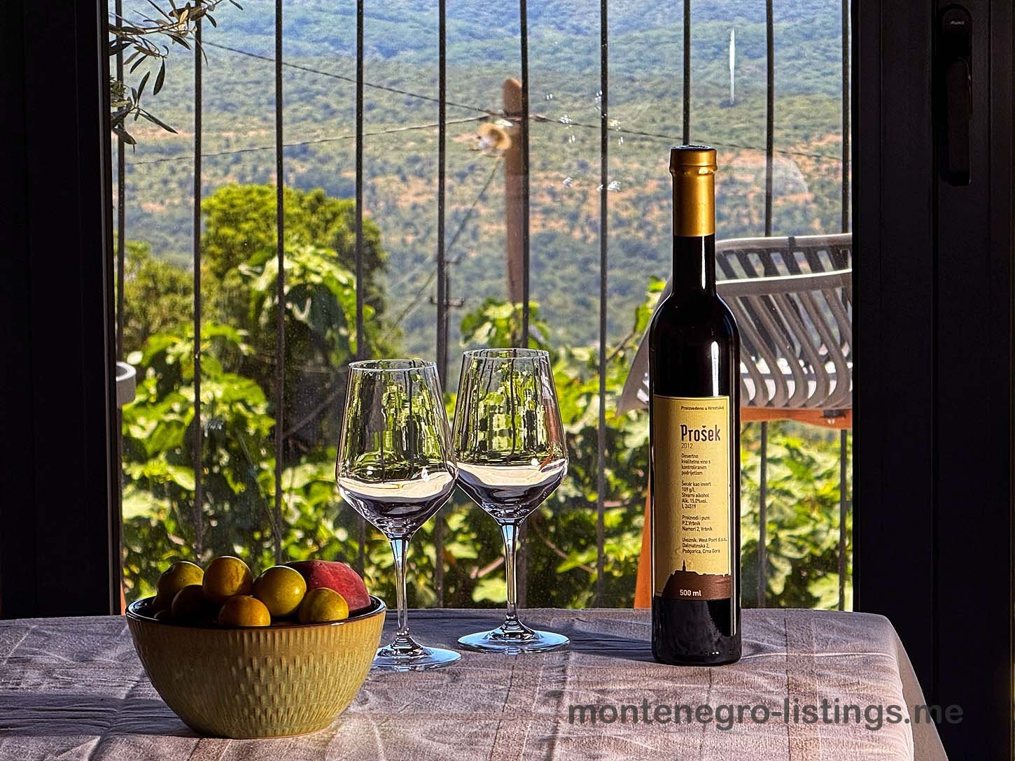 Pro&scaron;ek wine, glasses, and fresh fruit set by a window with green hill views&mdash;experience Ulcinj real estate luxury with immorenta.me.