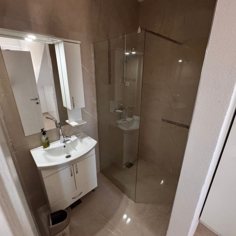 Modern bathroom in Ulcinj with beige tiles, glass shower, and white vanity. Invest with immorenta.me by Ekosphere doo.
