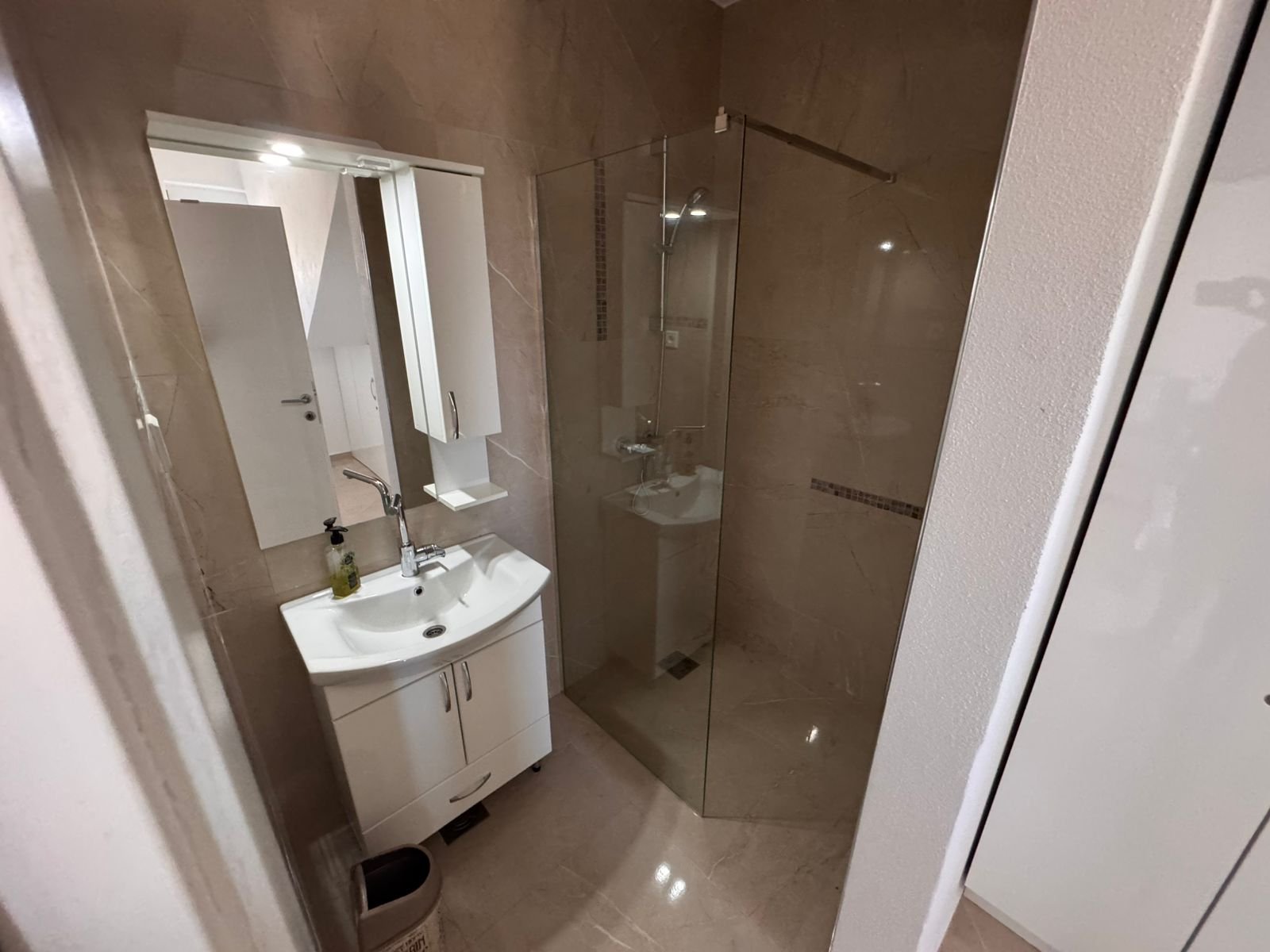 Modern bathroom in Ulcinj with beige tiles, glass shower, and white vanity. Invest with immorenta.me by Ekosphere doo.