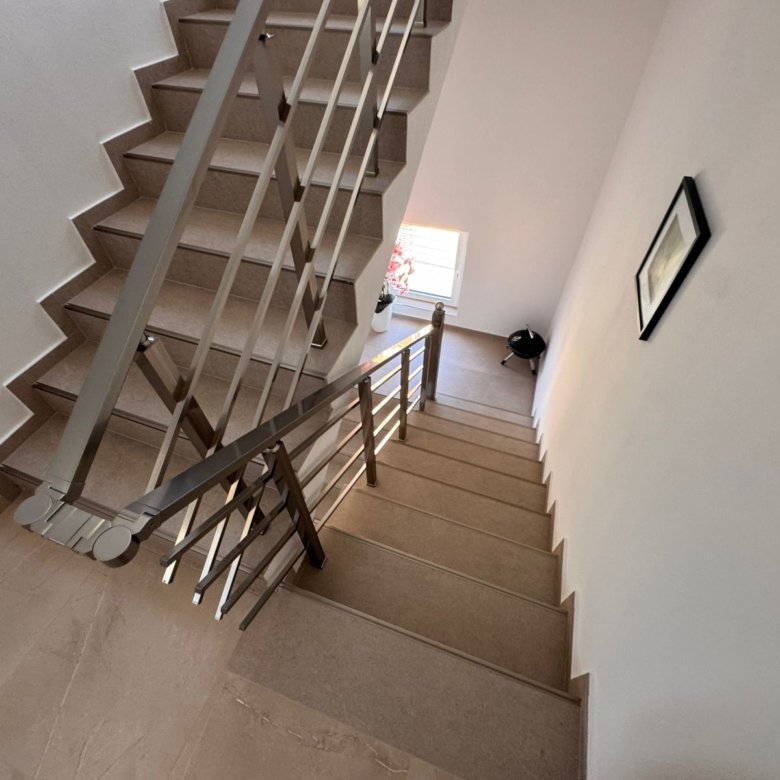 Elegant beige staircase with modern metal railings in Ulcinj property; bright landing with window, plant, and tasteful decor.