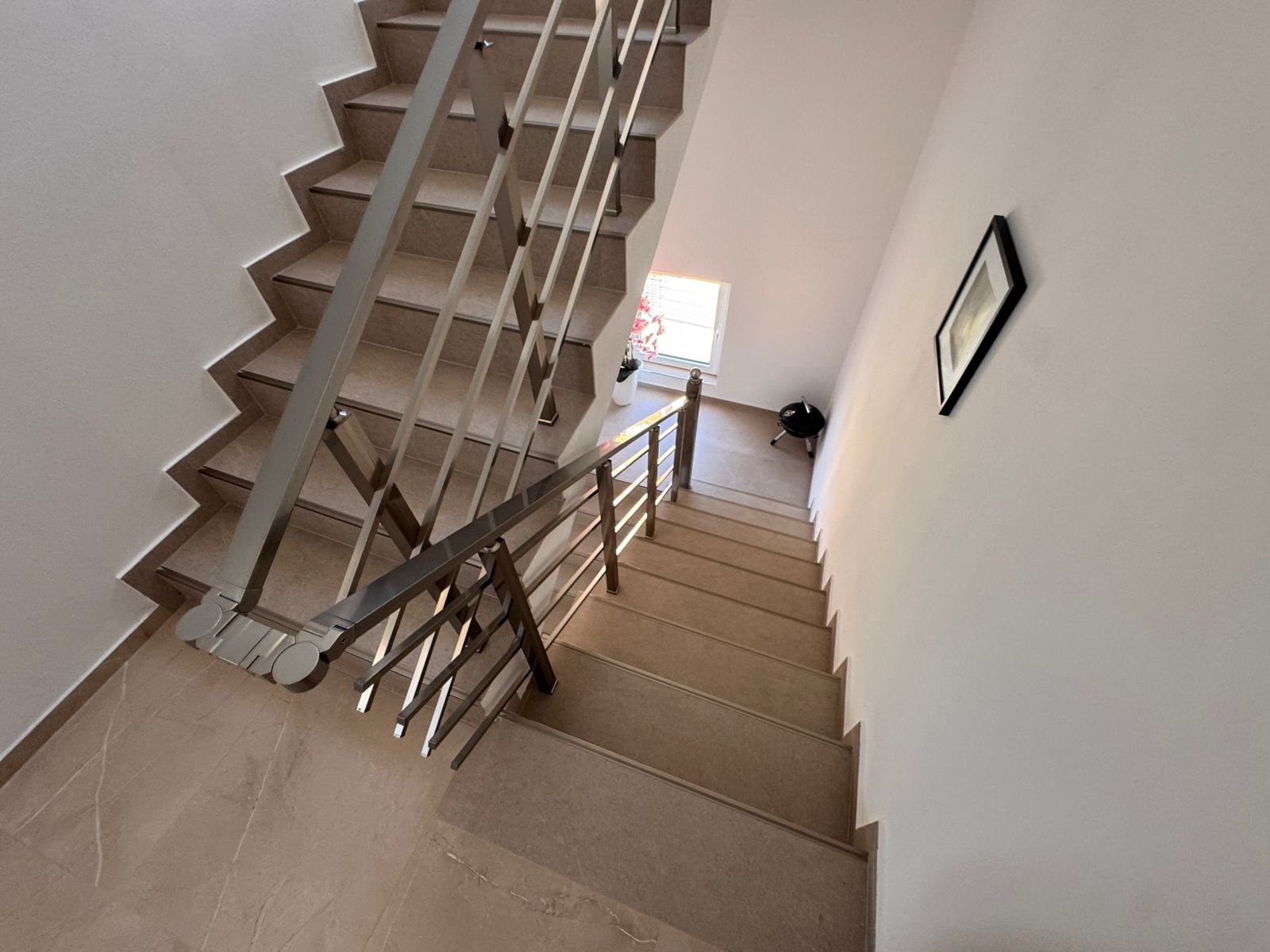 Elegant beige staircase with modern metal railings in Ulcinj property; bright landing with window, plant, and tasteful decor.