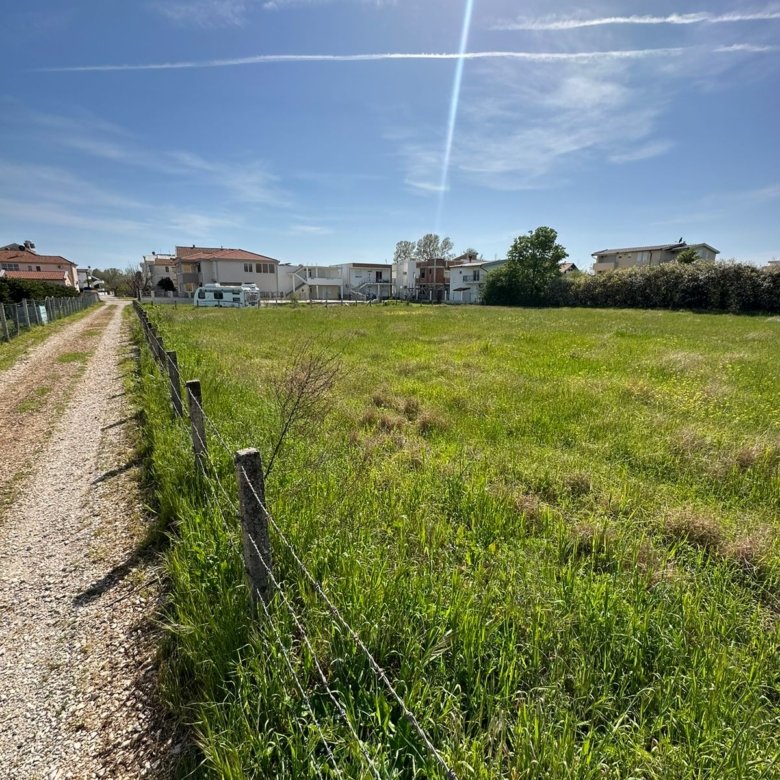Prime Ulcinj investment: Spacious grassy field with wire fence and gravel access, near sunny homes. Listed by ekosphere doo.