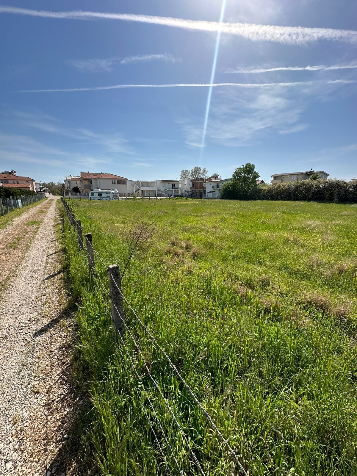 Prime Ulcinj investment: Spacious grassy field with wire fence and gravel access, near sunny homes. Listed by ekosphere doo.