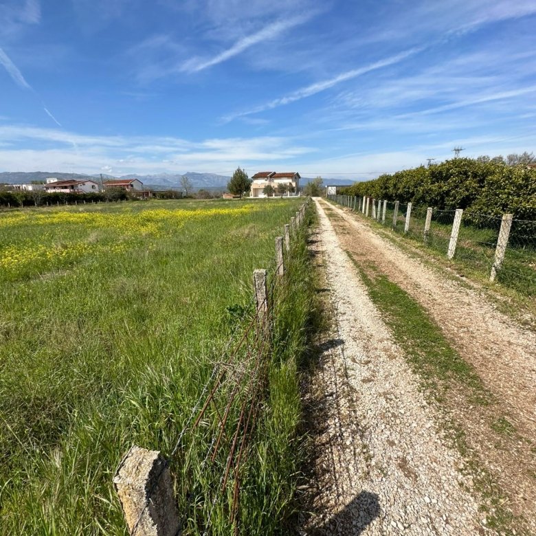 Prime Ulcinj land for sale: scenic gravel road, fenced grassy field with wildflowers, distant homes. Invest via immorenta.me.