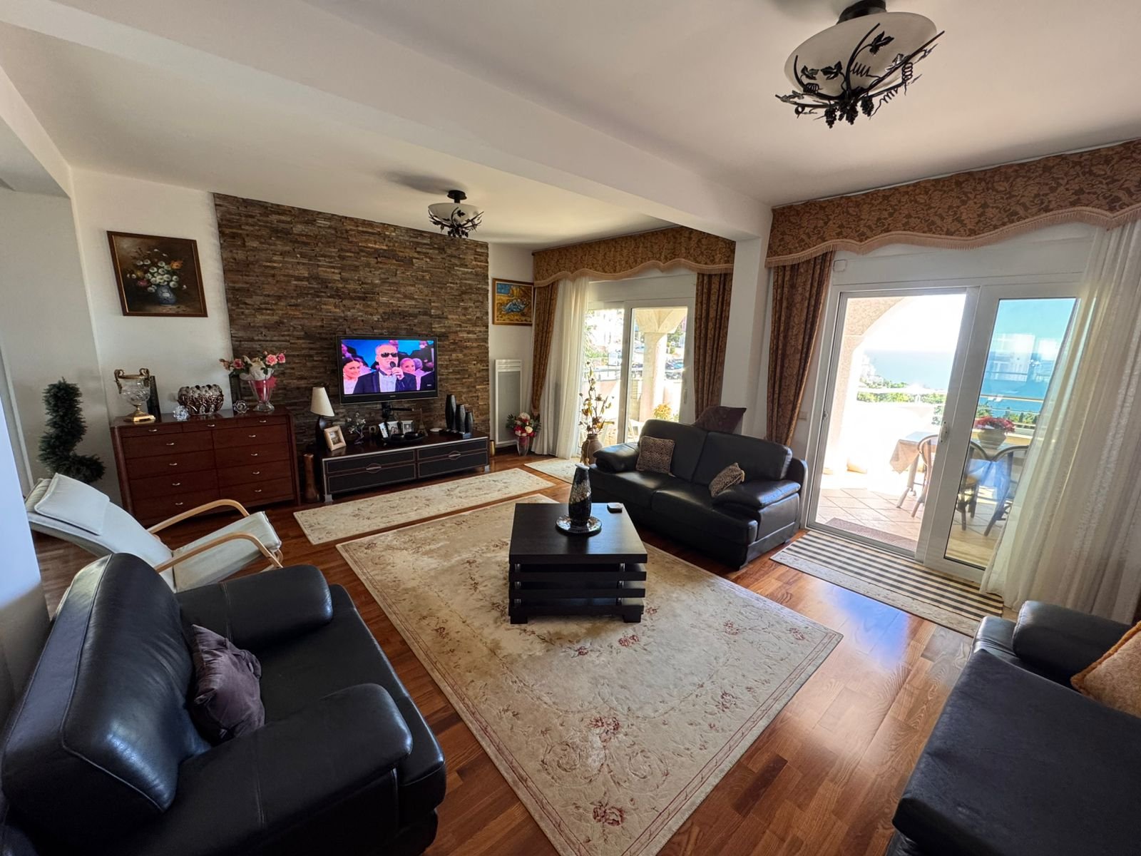 Elegant Ulcinj living room: black leather sofas, TV, stone wall, sea-view balcony. Invest via immorenta.me by ekosphere doo.
