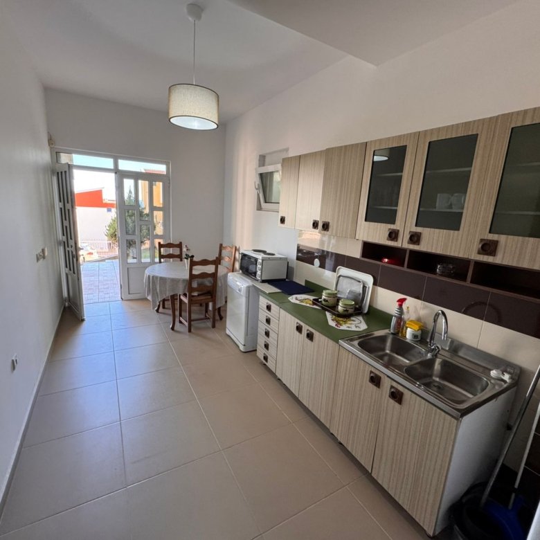 Modern Ulcinj kitchen with bright cabinets, double sink, dining area, and glass door to outdoor space&mdash;exclusive at immorenta.me.