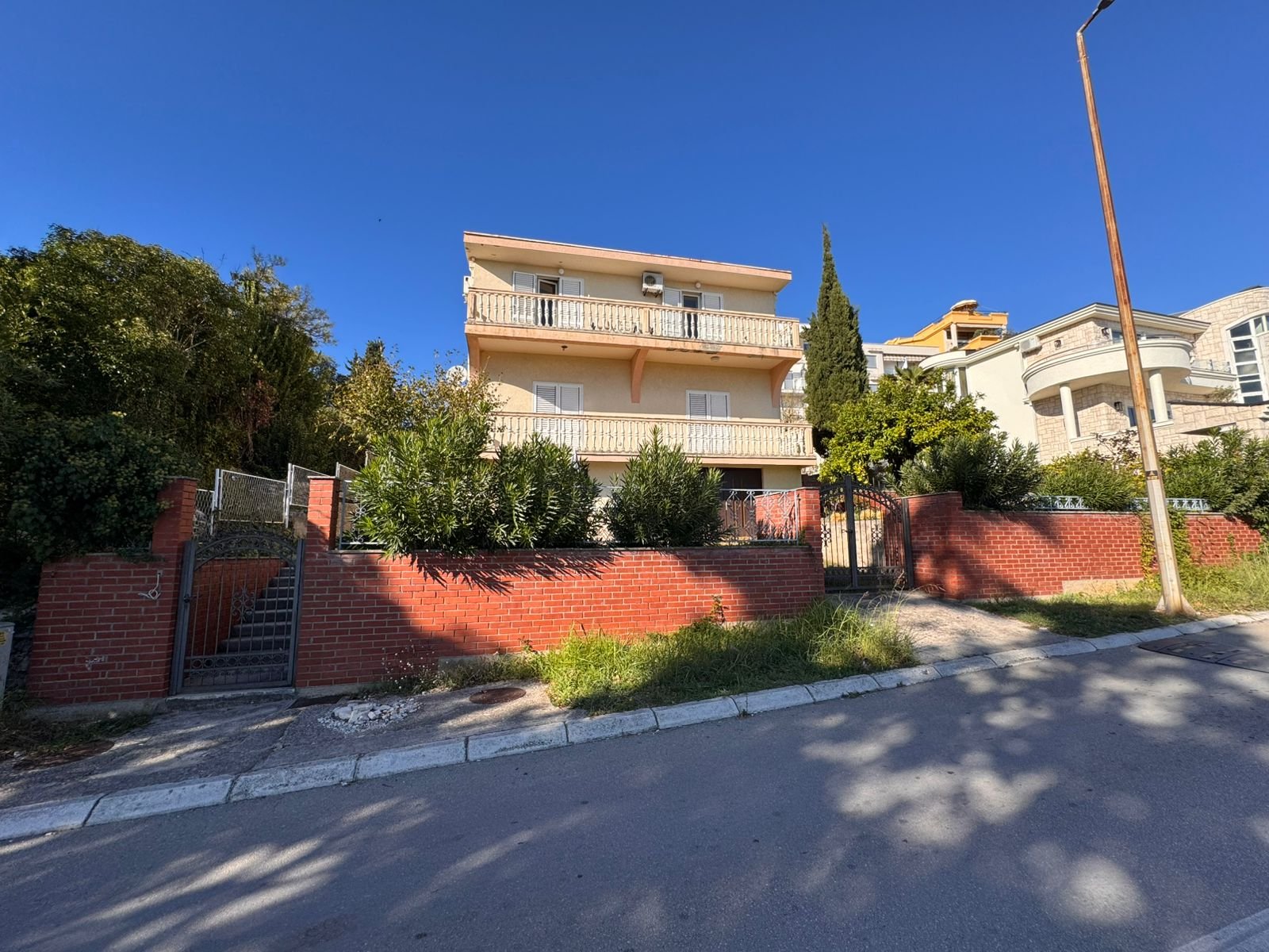 Stunning three-story yellow home with balconies, brick fence, and lush greenery in Ulcinj. Exclusive at immorenta.me by ekosphere doo.