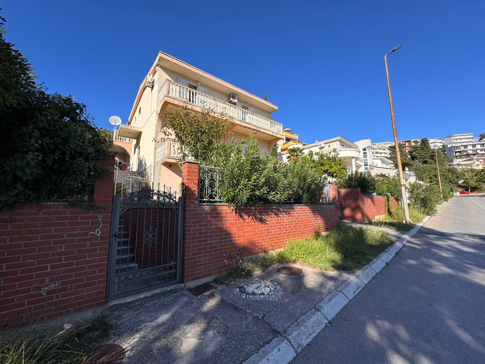 Modern two-story beige home with balconies, red brick fence, lush greenery, and gated entrance in Ulcinj, Montenegro.