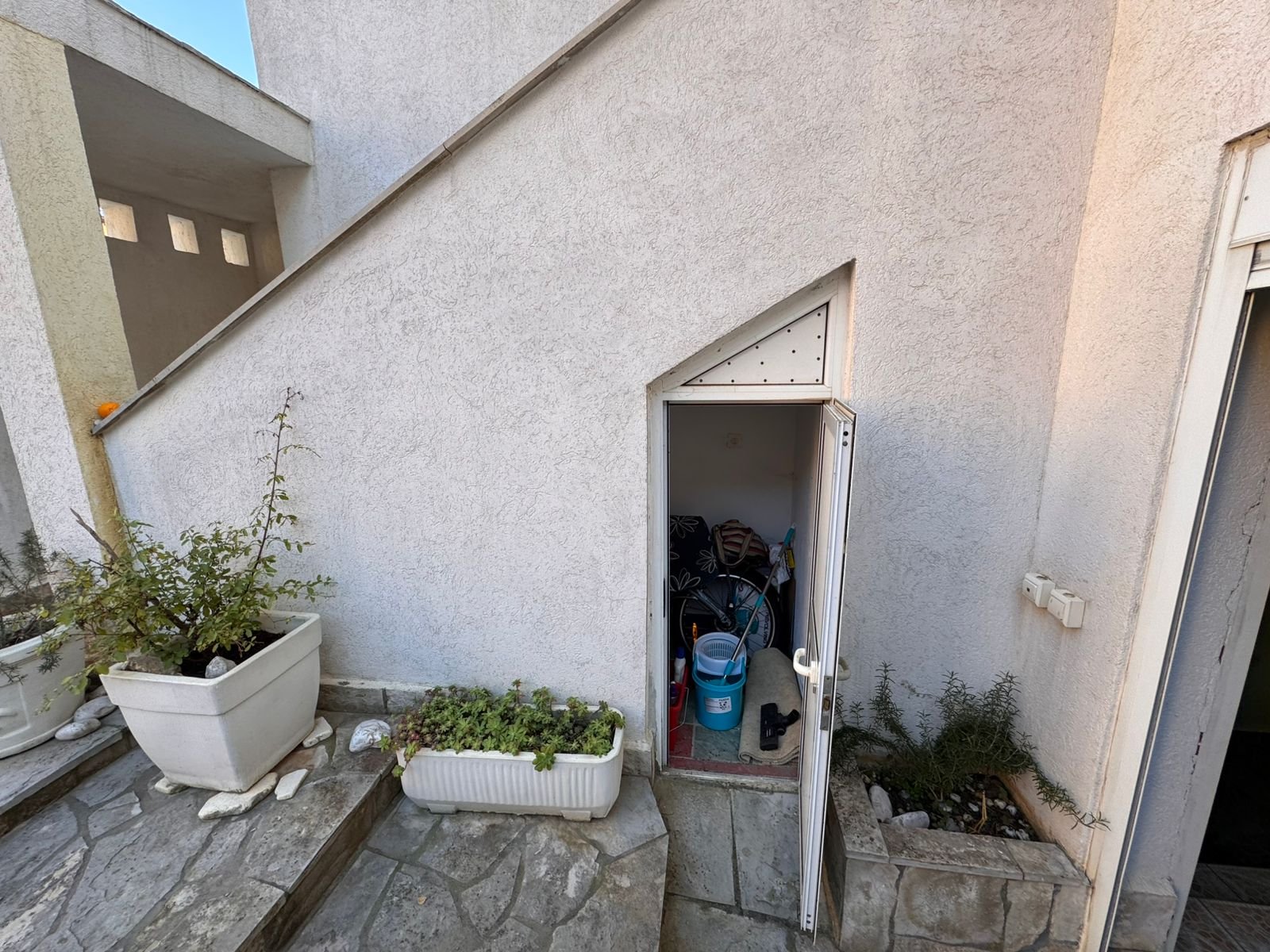Spacious white storage unit under stairs, ideal for Ulcinj Montenegro property investors. Includes organized tools, garden access, and entry plants. Montenegro-listings.me by ekosphere doo.