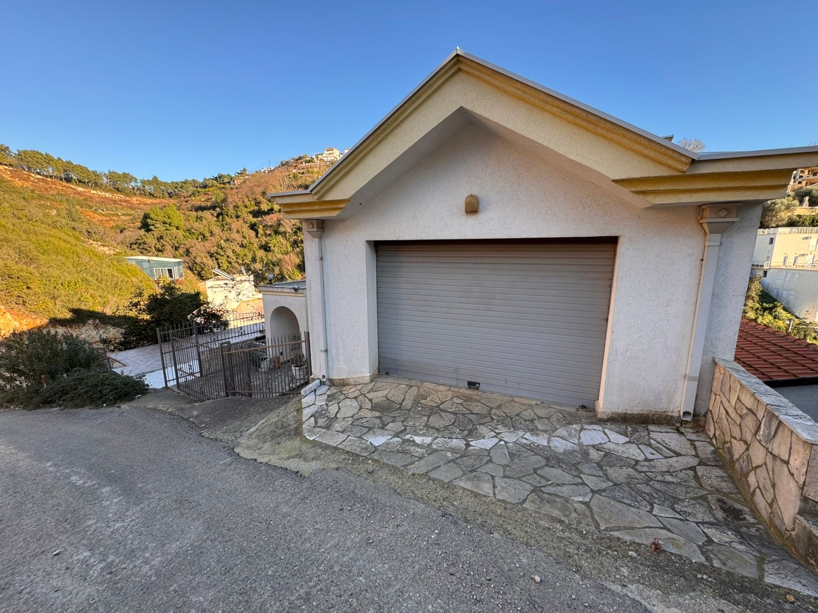 Modern white garage with secure gray door on a sloped lot, stone-paved drive, scenic Ulcinj views&mdash;exclusive at immorenta.me.