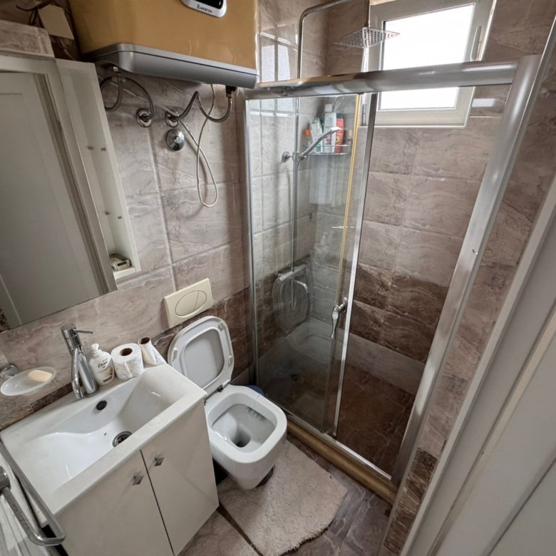 Modern bathroom in Ulcinj with beige tiles, glass shower, bright window, and quality fixtures. Exclusive at immorenta.me.