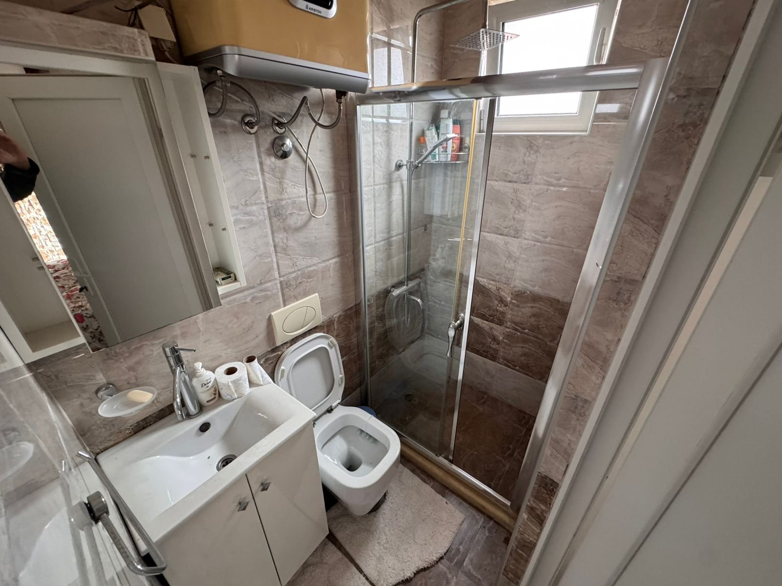 Modern bathroom in Ulcinj with beige tiles, glass shower, bright window, and quality fixtures. Exclusive at immorenta.me.