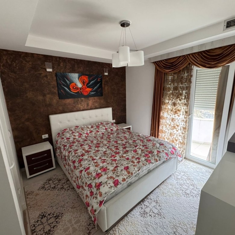 Modern Ulcinj bedroom with white bed, floral bedding, elegant nightstands, accent wall art, and large sunlit windows&mdash;exclusive at immorenta.me by ekosphere doo.