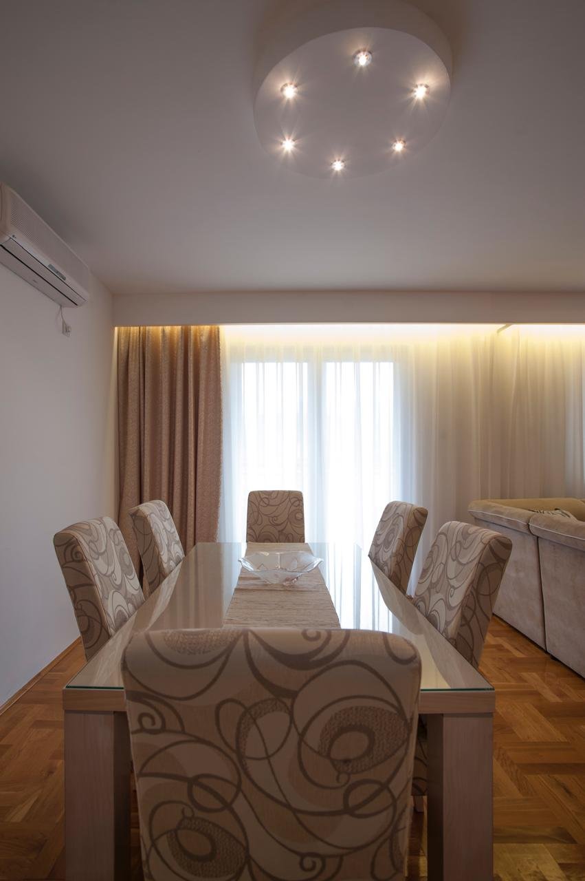 Elegant Ulcinj dining area: glass table, designer chairs, sunlit windows, chic lighting, hardwood floors. Invest via immorenta.me by ekosphere doo.