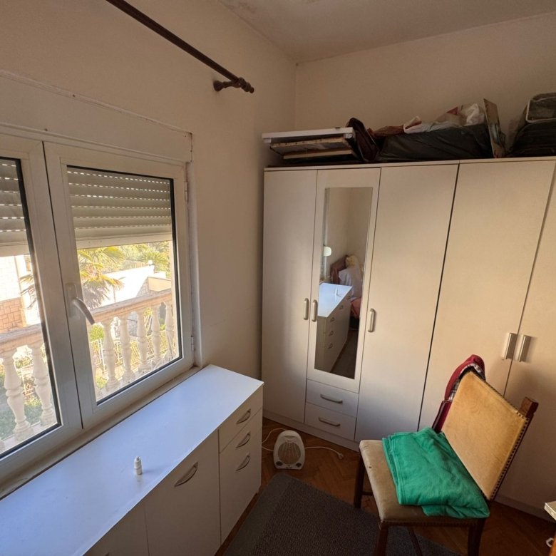 Bright furnished room in Ulcinj with white cabinets, mirrored wardrobe, cozy chair, sunlit window&mdash;prime Montenegro investment. Montenegro-listings.me by ekosphere doo.