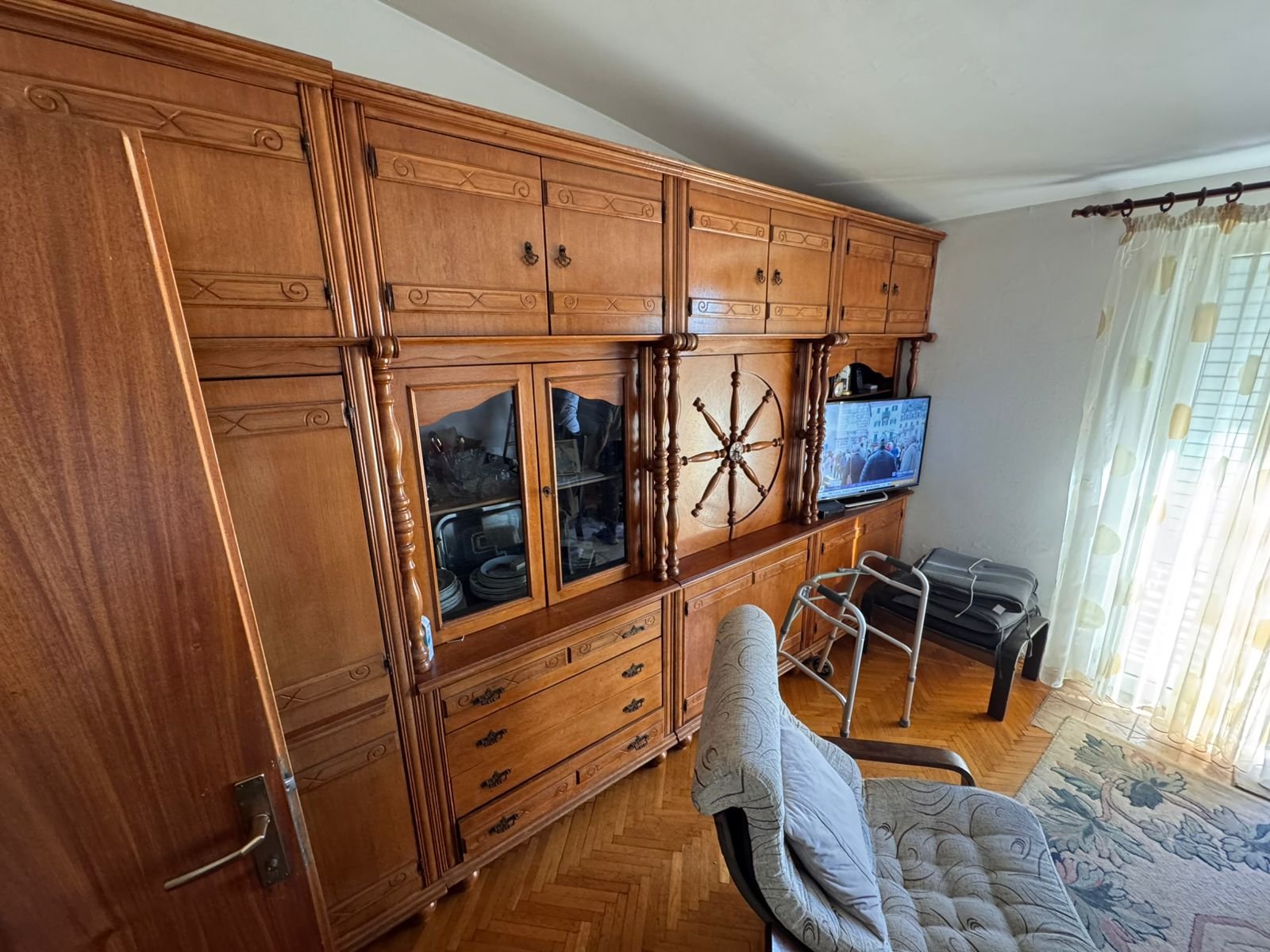 Elegant wooden wall unit with glass cabinets, TV, and cozy seating by a bright window&mdash;ideal Ulcinj real estate investment.