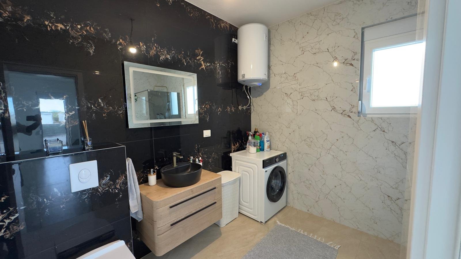 Luxurious Ulcinj bathroom: marble & black accents, wood vanity, washer, window. Prime Montenegro real estate by ekosphere doo.
