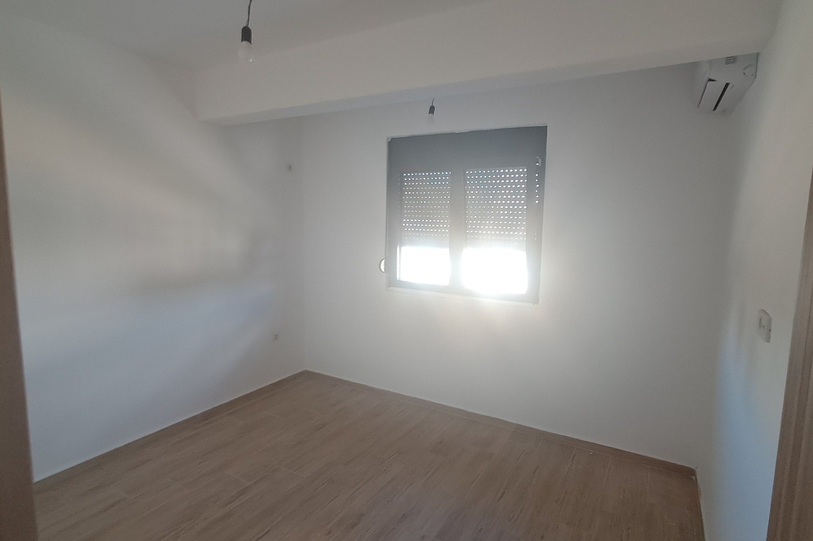 Bright, modern Ulcinj apartment with white walls, wood flooring, AC unit; sunlight through window. Invest via immorenta.me by ekosphere doo.
