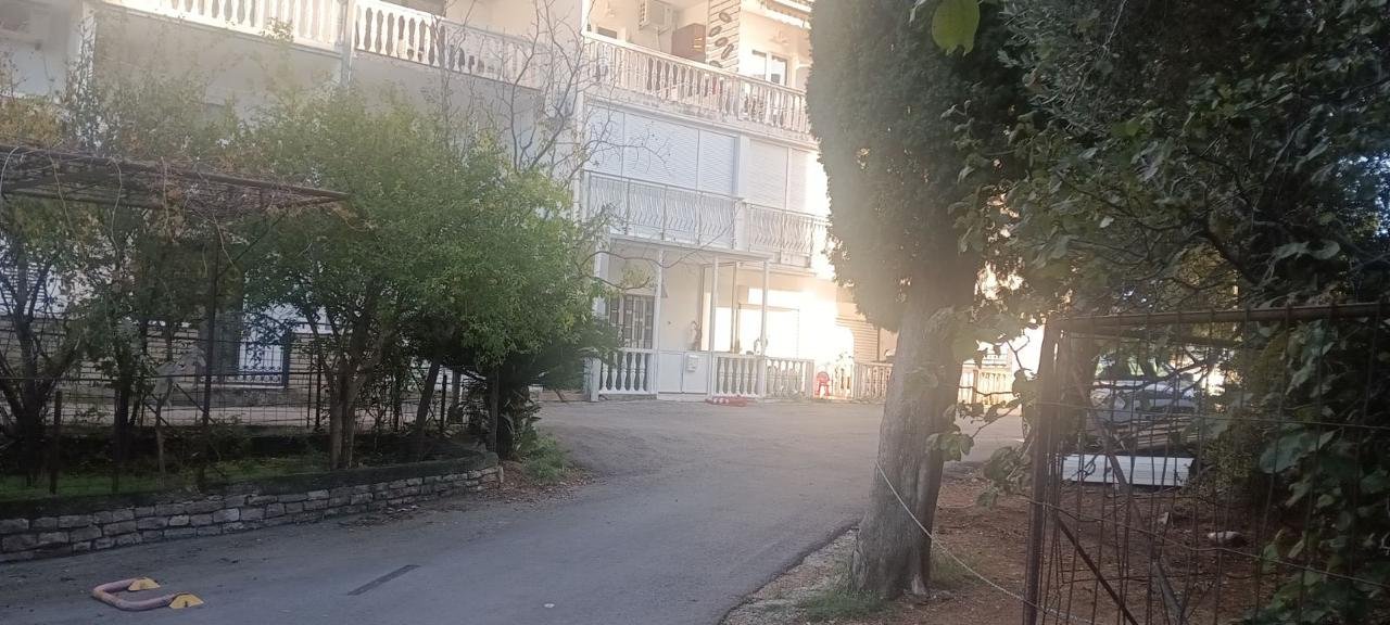 Charming Ulcinj property: white multi-story building with balconies, lush greenery, sunlit paved road&mdash;exclusive on immorenta.me by ekosphere doo.