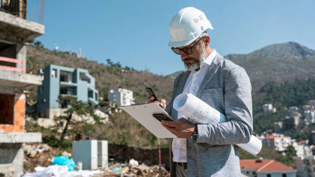 Ein Mann mit Schutzhelm und Brille hält Baupläne und ein Klemmbrett in der Hand, während er auf einer Baustelle mit teilweise fertiggestellten Gebäuden und Hügeln im Hintergrund auf sein Handy schaut.