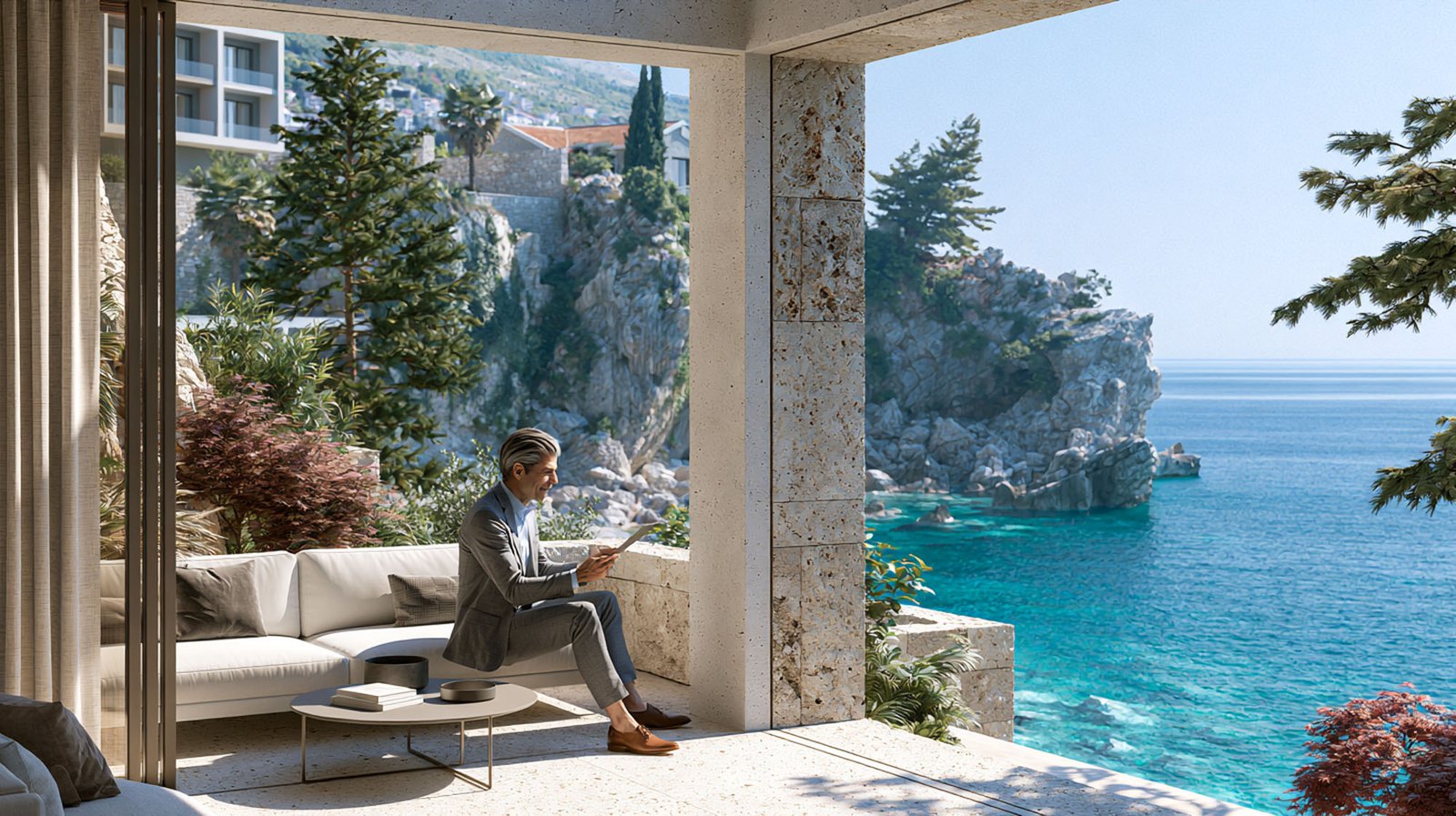 Eine Person im Anzug sitzt auf einem weißen Outdoor-Sofa auf einer Terrasse mit Blick auf Felsklippen, das blaue Meer, Bäume und moderne Gebäude im Hintergrund.