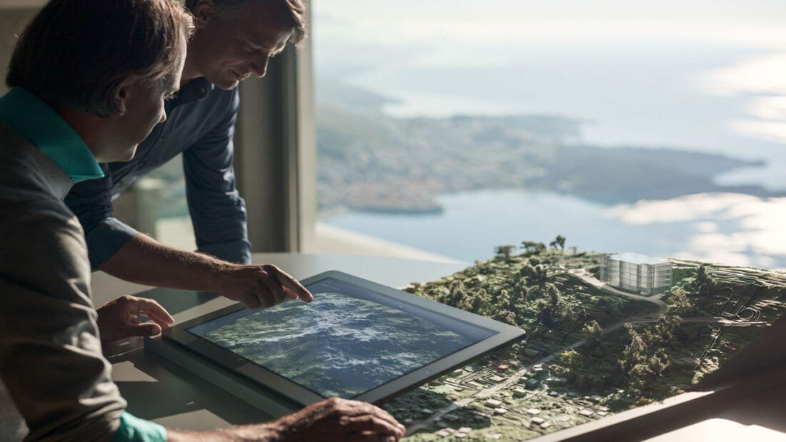 Zwei Personen betrachten eine digitale Karte auf einem Touchscreen neben einem Architekturmodell einer Landschaft, im Hintergrund ist durch ein großes Fenster der Blick auf die Küste und das Wasser zu sehen.