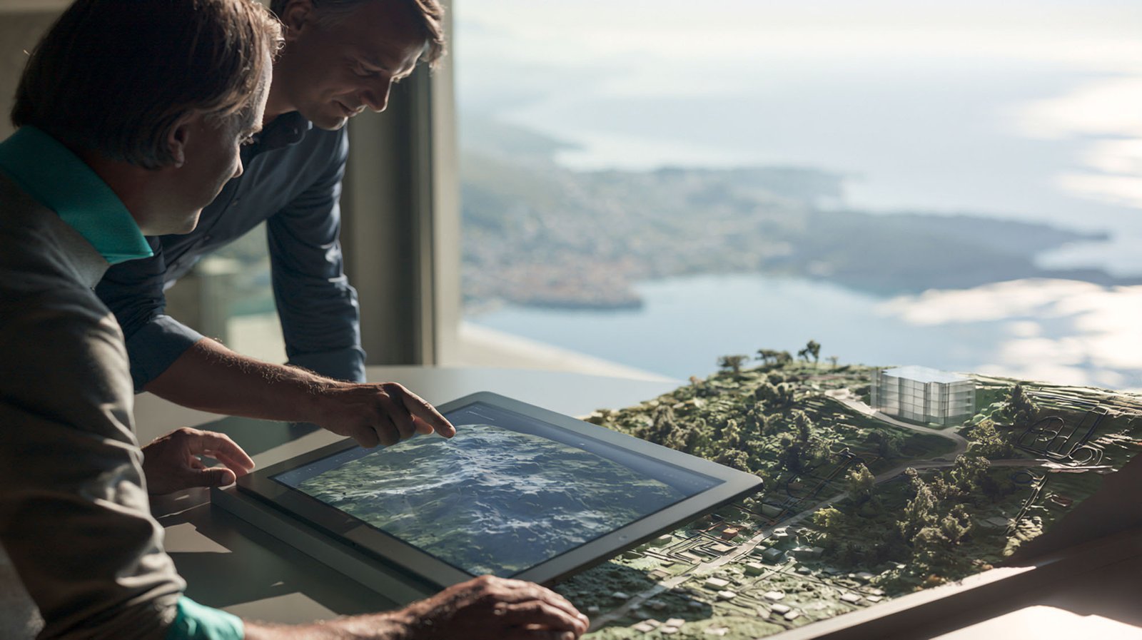 Zwei Personen betrachten eine digitale Karte auf einem Touchscreen neben einem Architekturmodell einer Landschaft, im Hintergrund ist durch ein großes Fenster der Blick auf die Küste und das Wasser zu sehen.